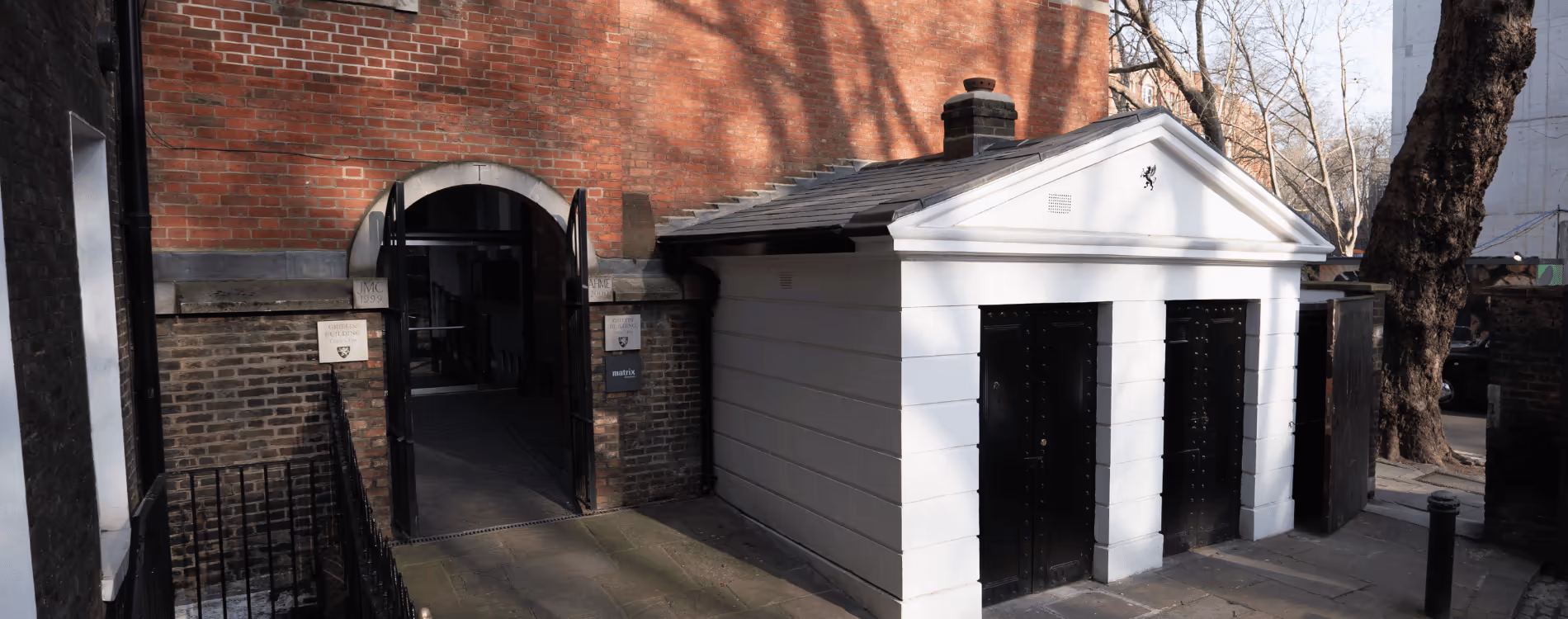 The exterior of a converted heritage gatehouse, painted white with black doors, standing next to the arched entrance of a larger historic red brick building.