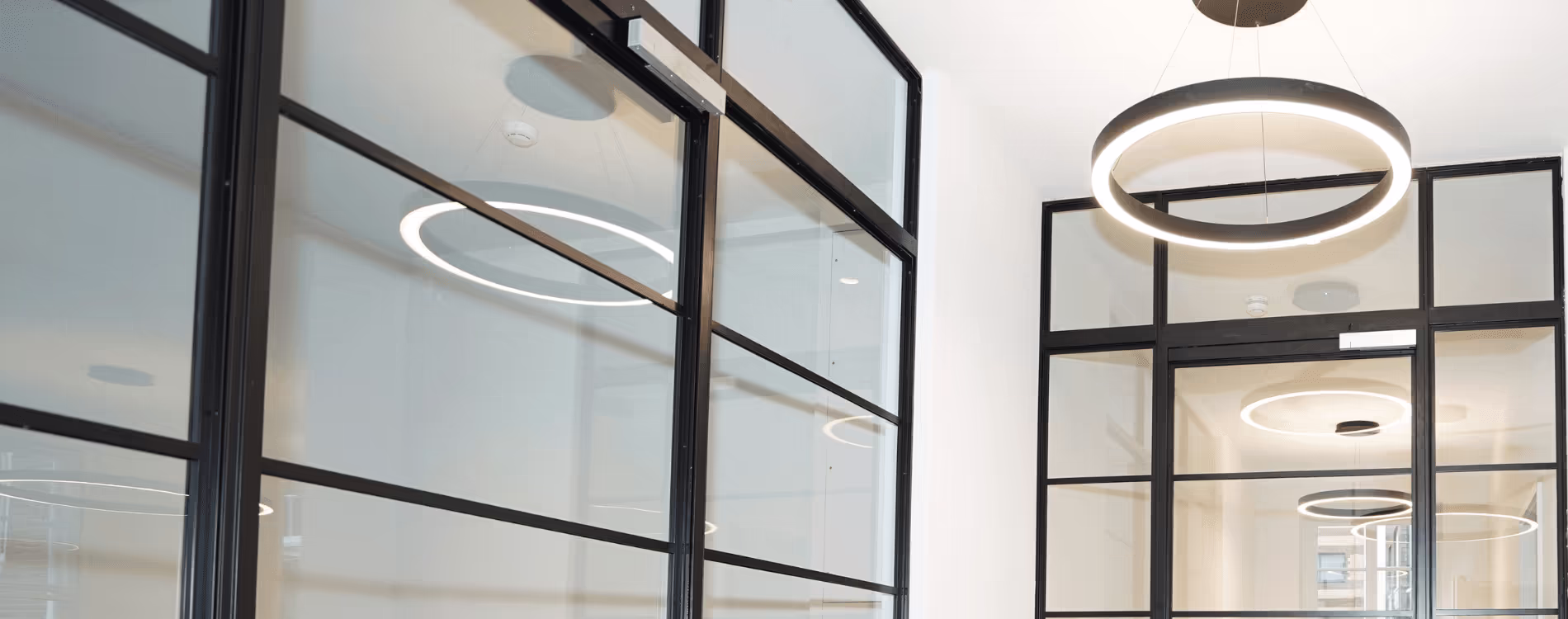 A modern refurbished office corridor featuring black-framed Crittall-style glass walls and contemporary circular LED pendant lights, which are reflected in the glass.